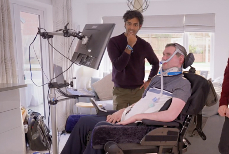 Ventilated man in a wheelchair looking at a screen, another man watches.